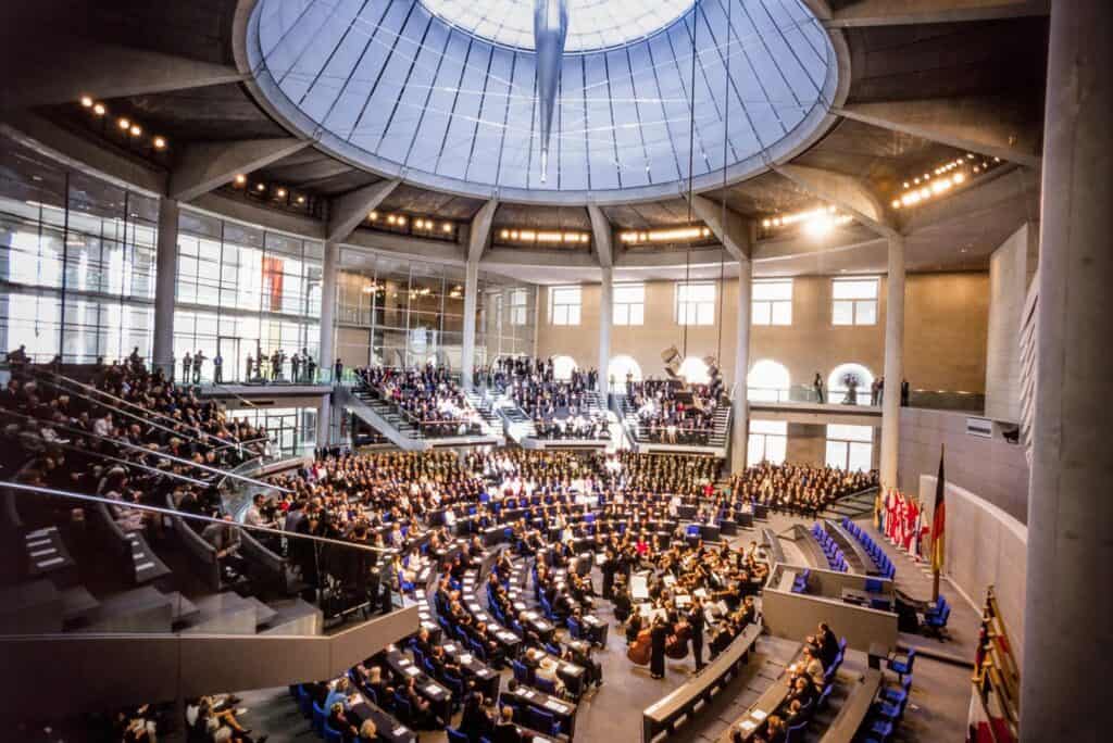 Plenary chamber of the Reichstag full of members and guests for a ceremony, tiers of blue seats beneath a round glazed ceiling and the tip of the mirrored cone, flags beside the stone walls