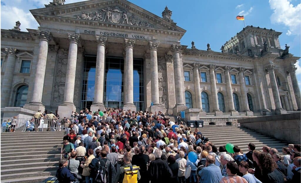Crowds of visitors queuing on the broad stone steps of the Reichstag's west portico beneath Corinthian columns and the pediment inscribed Dem Deutschen Volke