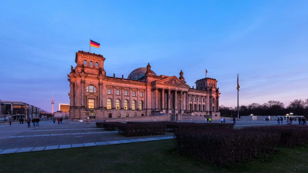 The Reichstag at sunset seen from the Platz der Republik, its west facade glowing pink beneath a violet sky, the Berlin TV Tower and the glass cupola on the skyline