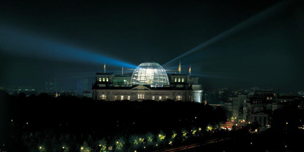 The Reichstag at night with its illuminated glass cupola glowing above the dark mass of the Tiergarten and searchlight beams sweeping across the sky