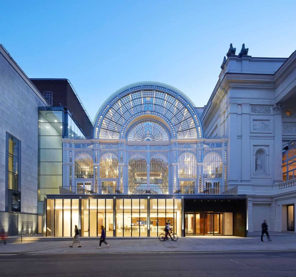 Bow Street front of the Royal Opera House at dusk, the arched iron and glass Floral Hall rising above a new glowing glazed pavilion at street level, with pedestrians and a cyclist passing and the stone-clad 1990s wing to the left