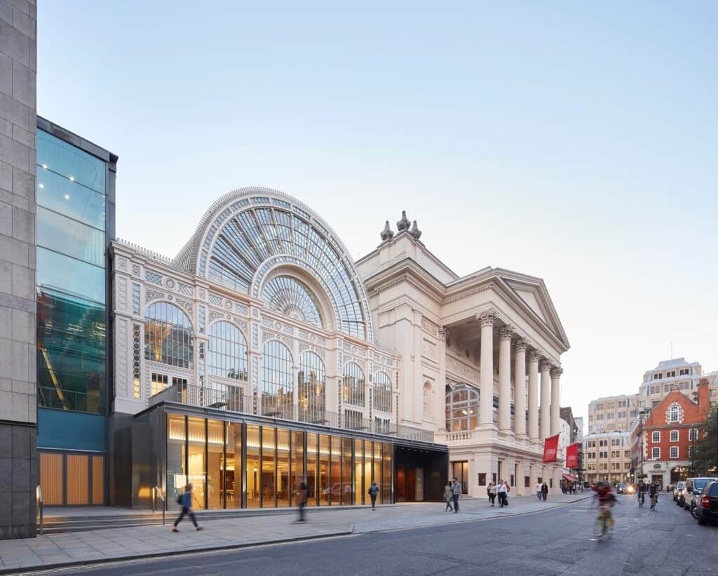 Angled view along Bow Street of the Royal Opera House in evening light, the restored Floral Hall and its glazed ground-floor pavilion leading to E.M. Barry's columned portico, with a red brick building and parked cars beyond