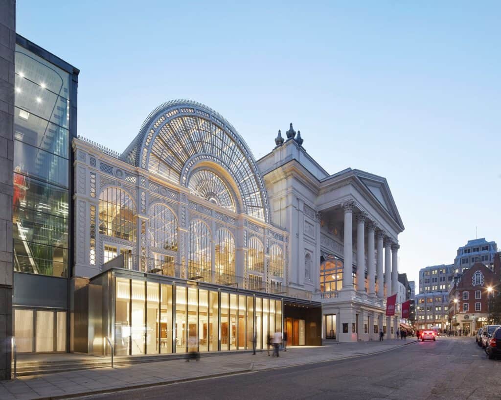 Royal Opera House on Bow Street at twilight, the warmly lit glass pavilion beneath the fan-shaped arch of the Floral Hall next to the pedimented Corinthian portico, a car's lights glowing down the street