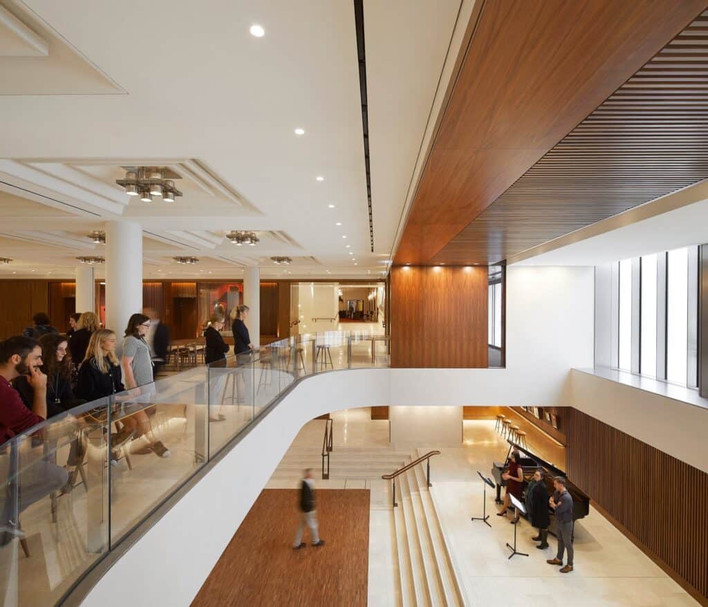 Visitors leaning on a curved glass balustrade overlooking the double-height foyer of the Royal Opera House, where singers rehearse beside a grand piano at the foot of pale stone steps beneath a walnut-clad soffit