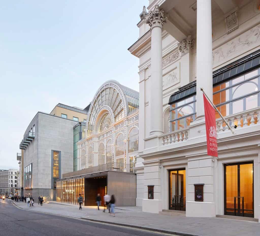 Corinthian columns and carved stone of E.M. Barry's portico on Bow Street with a red Royal Opera House banner, the Floral Hall arch and its new glazed pavilion receding along the street behind