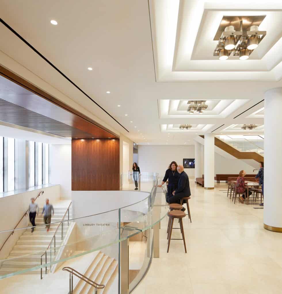 Enlarged entrance foyer of the Royal Opera House with a sweeping glass balustrade around the stair down to the Linbury Theatre, visitors at high tables with walnut stools under square light fittings set in a coffered ceiling