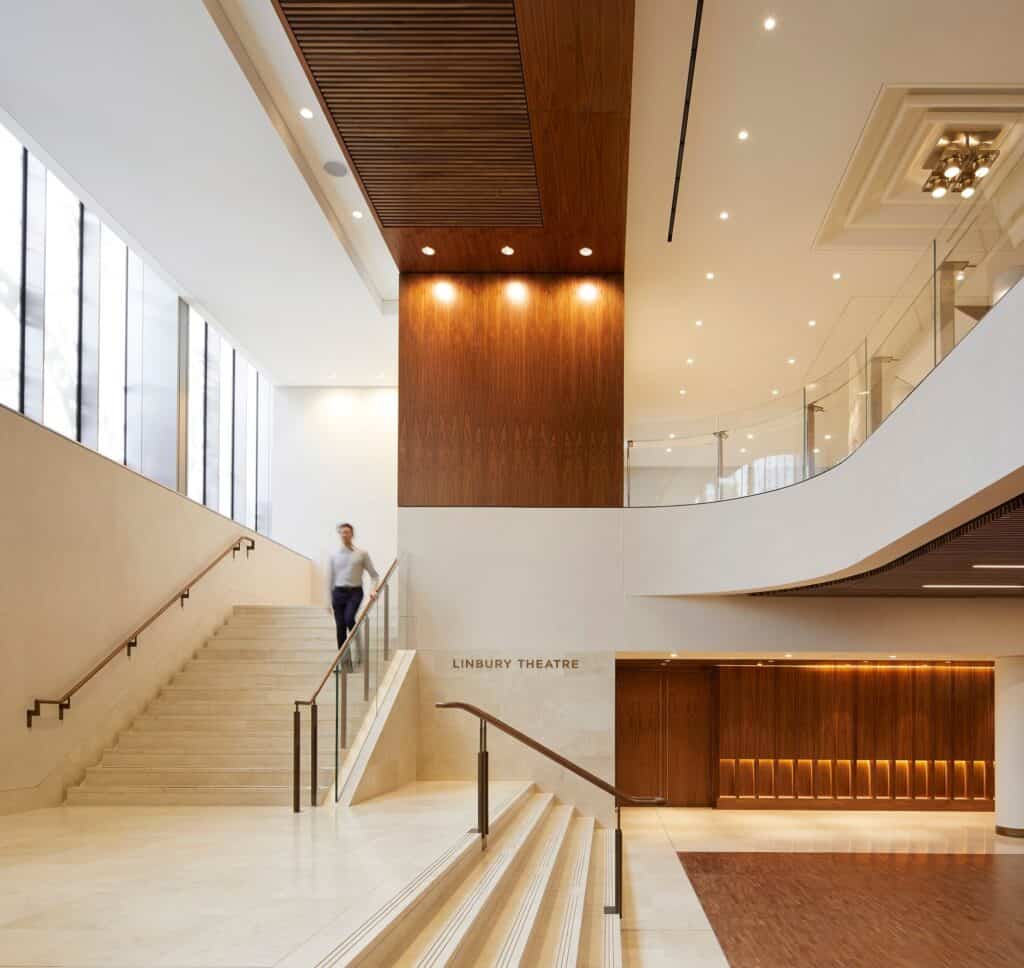Double-height space at the Royal Opera House, pale stone stairs with bronze handrails descending to the walnut-panelled Linbury Theatre entrance beneath a curving white balcony and a slatted timber soffit