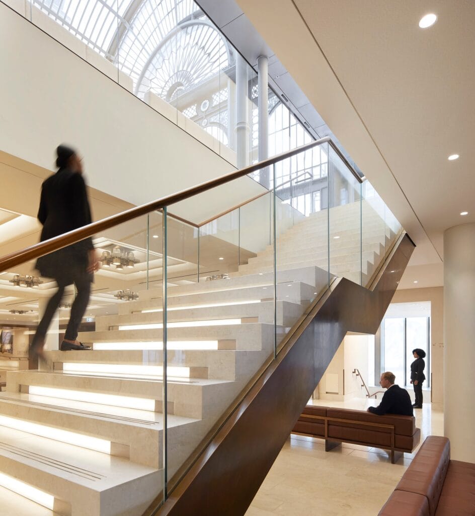 A visitor climbing a glass-balustraded stone stair with lit treads and a walnut handrail towards the Paul Hamlyn Hall, the arched glazing of the Floral Hall visible above and a man seated on a leather bench below