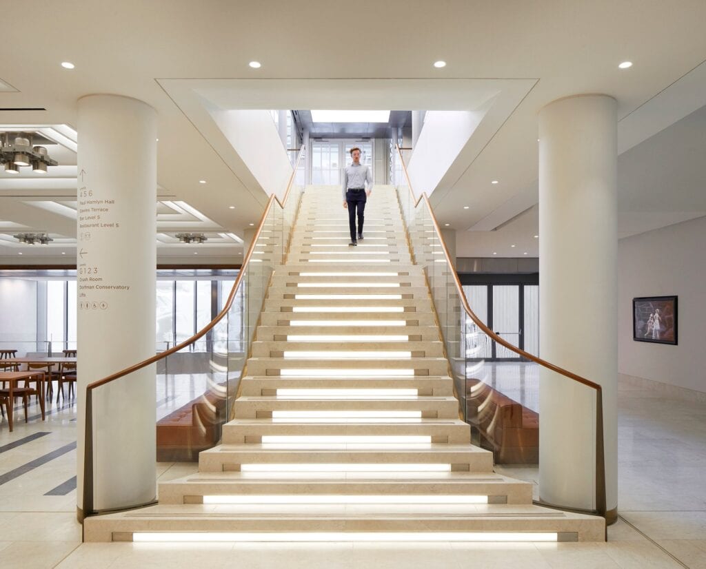 Broad stone staircase with illuminated treads and glass balustrades rising between two white columns at the Royal Opera House, a man walking down it past foyer tables and wayfinding signs