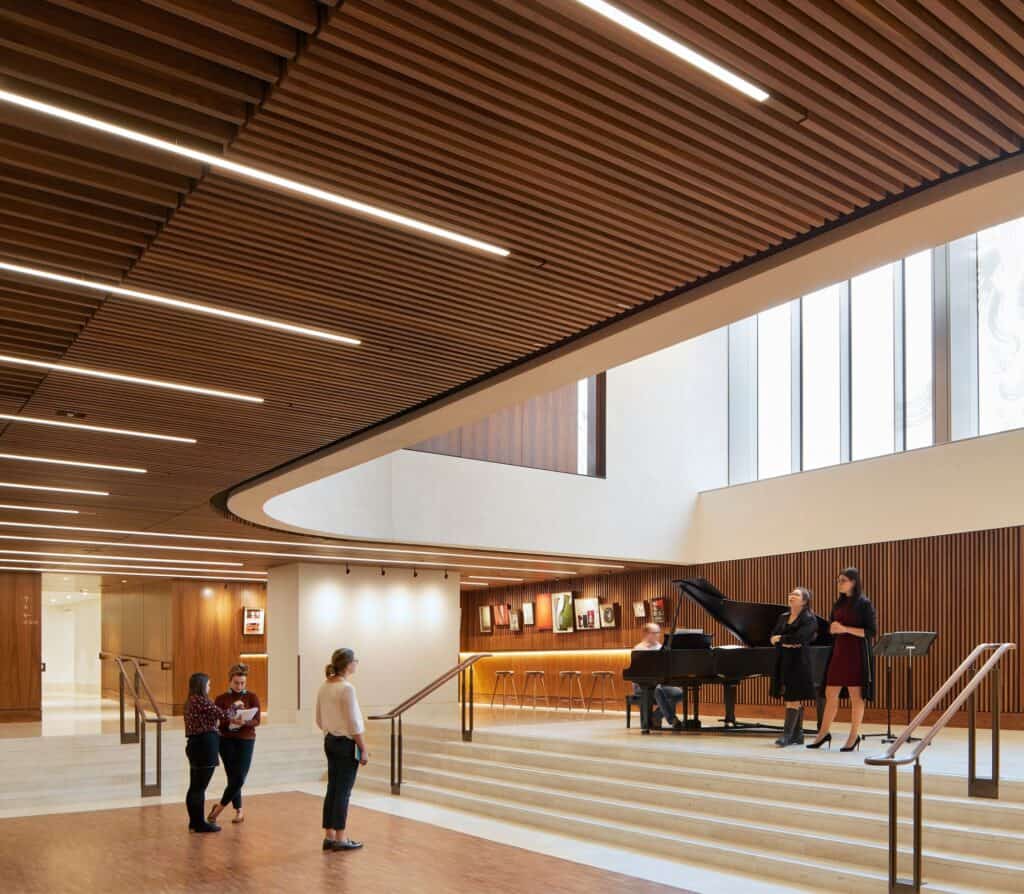 Linbury Foyer at the Royal Opera House with a slatted timber ceiling cut by linear lights, singers and a pianist at a black grand piano on a raised stone platform, a curved opening and tall windows above