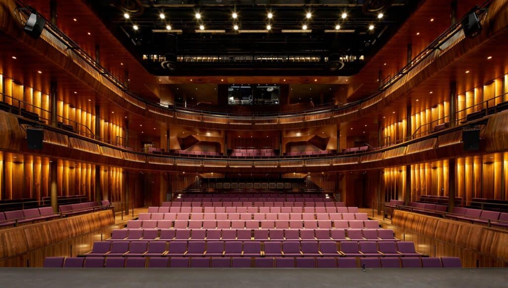 The Linbury Theatre at the Royal Opera House seen from the stage, rows of pink-purple seats rising to two tiers of curving galleries lined in glowing American black walnut
