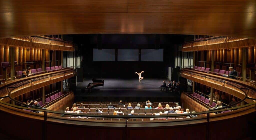 Dancer performing on the stage of the Linbury Theatre beside a grand piano, watched by an audience in the stalls and in the walnut-fronted side galleries