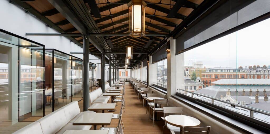 Long restaurant on the Amphitheatre level of the Royal Opera House, white tables and banquettes along tall windows looking over the Covent Garden market roofs beneath dark timber trusses and hanging lanterns