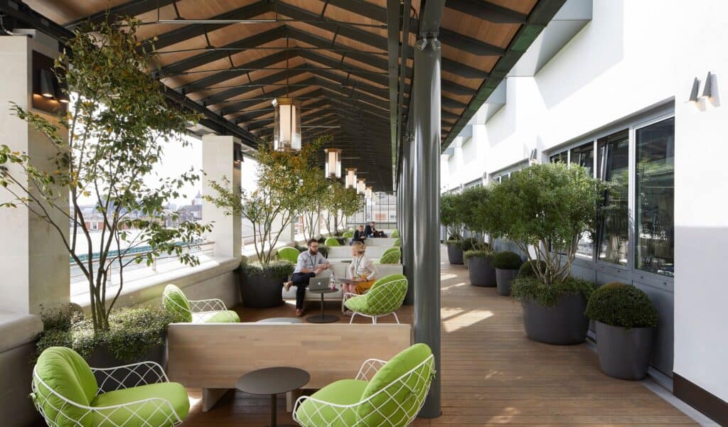 Amphitheatre Terrace at the Royal Opera House under a pitched timber roof, lime green wire armchairs, potted trees and clipped shrubs in grey planters, with two people talking on a bench