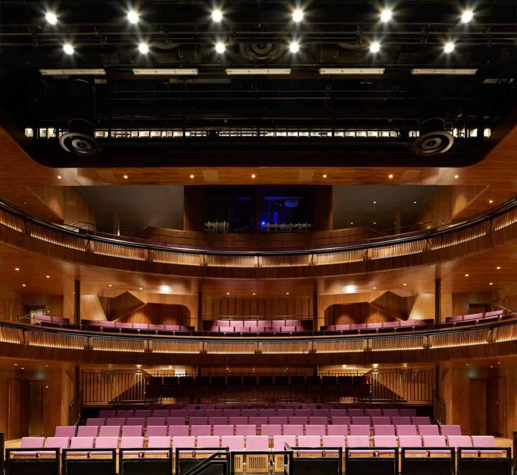 The Linbury Theatre from the stage, walnut-lined balconies stacked beneath a black technical grid of lights, pink seats in the stalls and a blue-lit control room at the rear