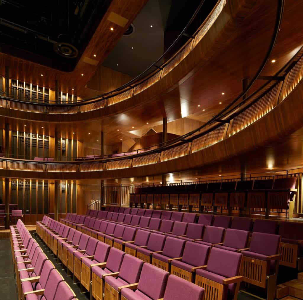 Side view across the Linbury Theatre, pink upholstered seats with timber ends in the stalls below curving walnut galleries with backlit slatted balustrades and slim brass handrails