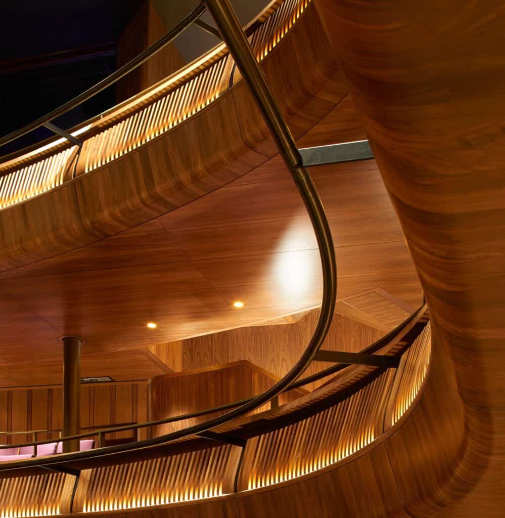 Looking up at the sculpted curves of the walnut-clad galleries in the Linbury Theatre, their backlit slatted fronts and a sinuous brass handrail glowing against the dark