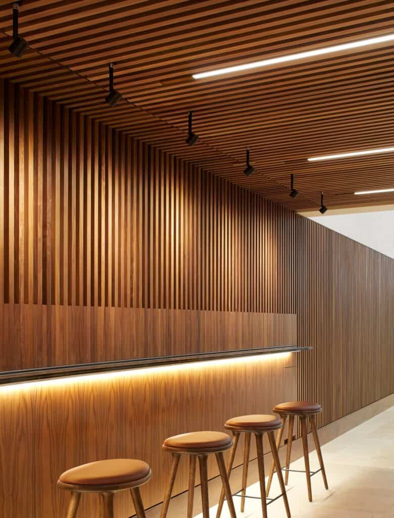 Walnut bar at the Royal Opera House with a slim illuminated counter set against vertical timber slats, four leather-topped wooden stools and a slatted ceiling with track spotlights