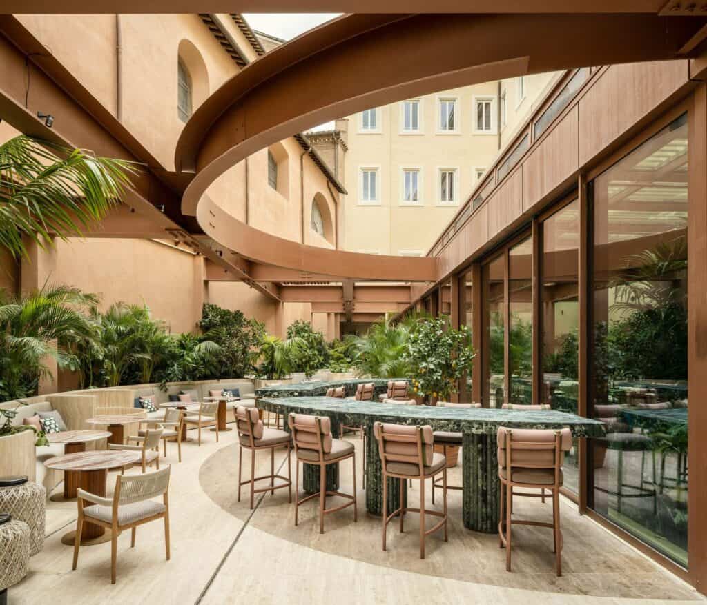 Courtyard of Six Senses Rome in Palazzo Salviati Cesi Mellini, a curved green marble bar with high leather stools on travertine paving beneath an elliptical rust-red steel ring, with palms and the ochre palazzo walls and windows rising behind
