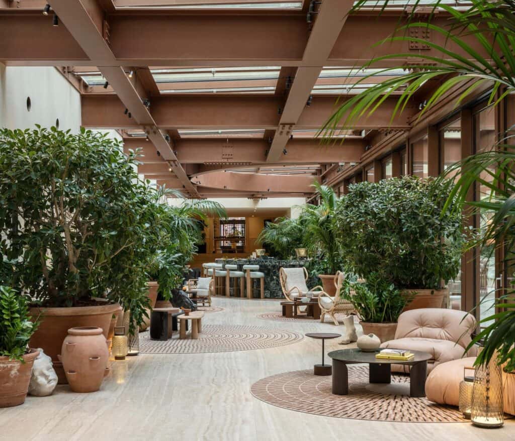 Glass-roofed lobby gallery at Six Senses Rome, rust-red steel beams above travertine floors, potted citrus trees and terracotta amphorae, round woven rugs and low armchairs leading to a bar at the far end