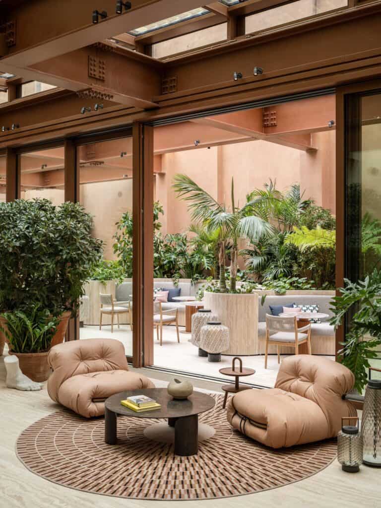Lobby lounge at Six Senses Rome with two pink leather Soriana armchairs and a round black table on a circular patterned rug, looking through a tall glazed wall to the planted courtyard