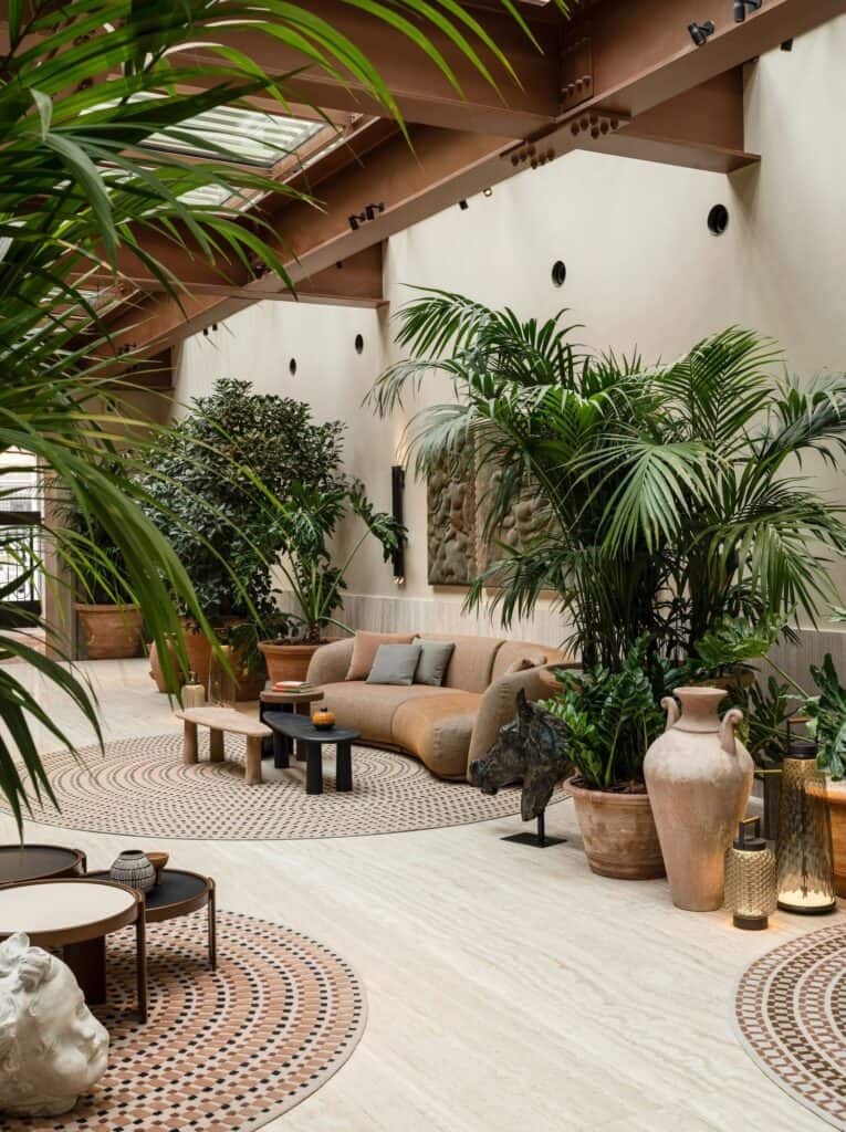 Corner of the Six Senses Rome lobby with a curved pink sofa and low tables on round mosaic-patterned rugs, tall palms, terracotta amphorae and a white stone fragment on the travertine floor
