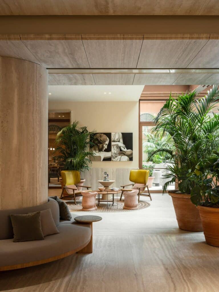 View past a thick travertine-clad column wrapped by a curved grey upholstered bench into a lounge with mustard yellow armchairs, small terracotta tables, a large black and white artwork and potted palms