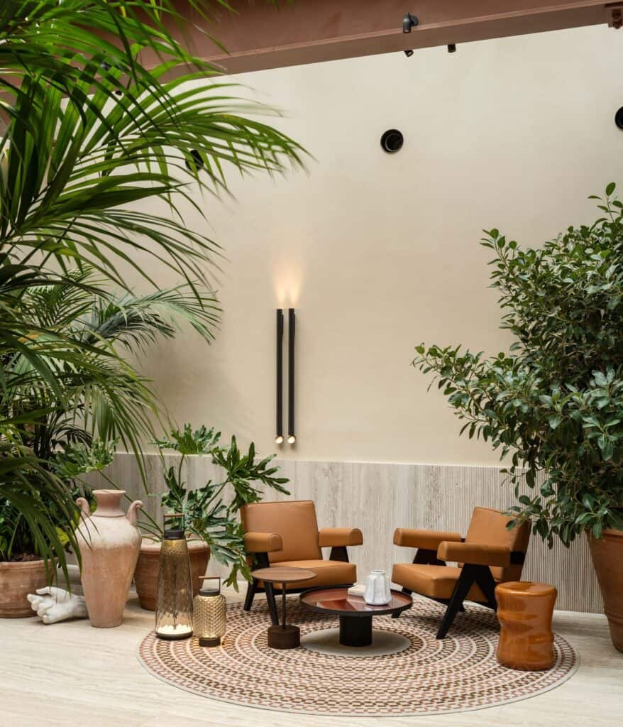 Seating nook at Six Senses Rome with a pair of tan leather armchairs on a round rug against a pale plaster wall with a slim black wall light, framed by palms, a ficus and terracotta urns
