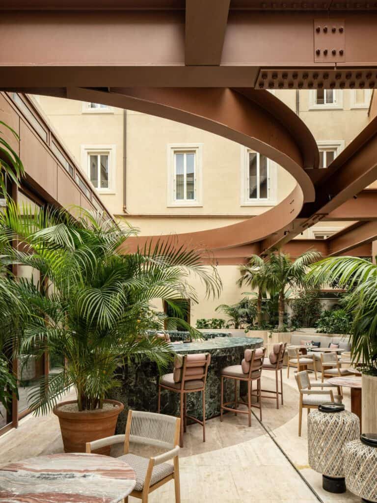 Courtyard of Palazzo Salviati Cesi Mellini seen beneath a curving rust-red steel ring, the green marble bar and leather stools among potted palms with the pale yellow palazzo façade and its windows above