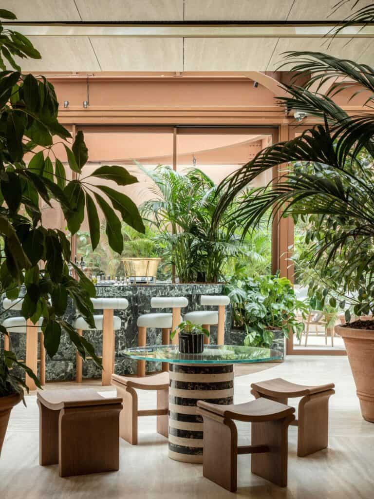 Bar corner at Six Senses Rome with wooden stools around a glass-topped table on a striped black and white marble base, white bar stools at a green marble counter and leafy palms under the skylight