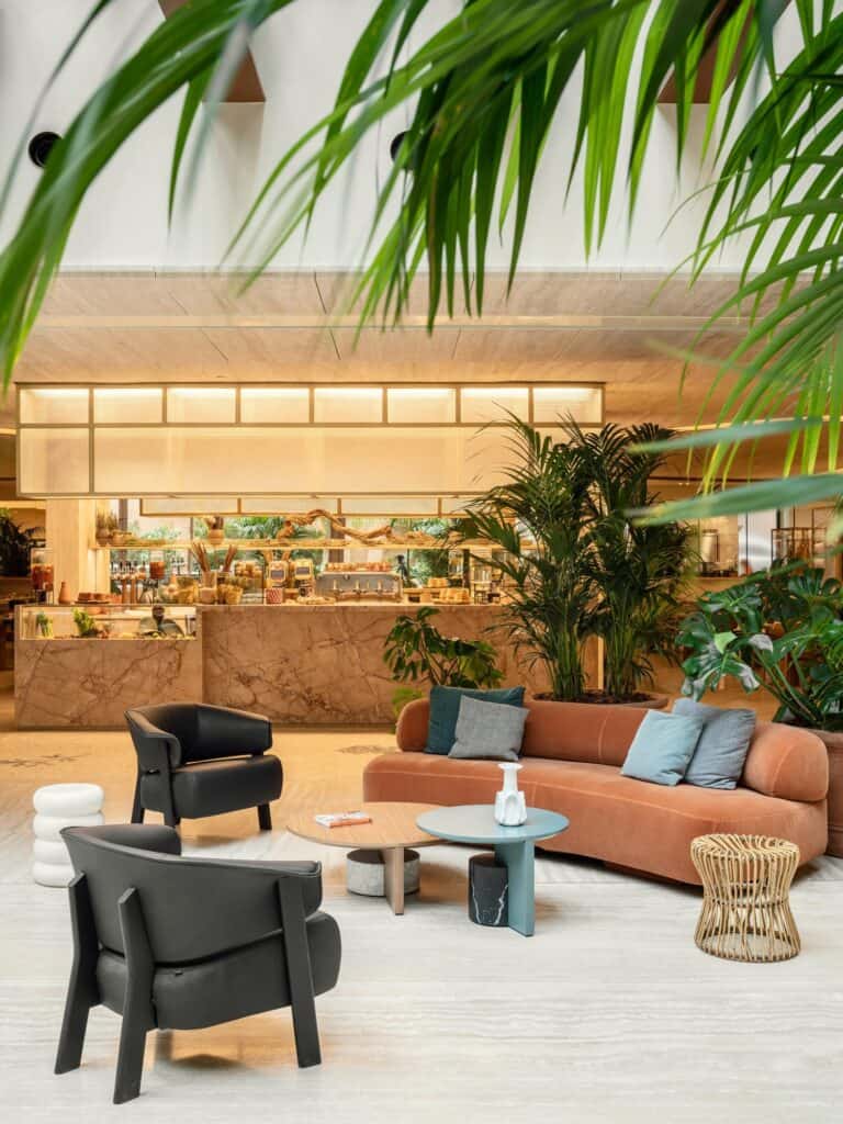Bivium restaurant-café at Six Senses Rome, a terracotta curved sofa with blue cushions, black armchairs and a teal side table in front of the open kitchen counter, framed by palm fronds