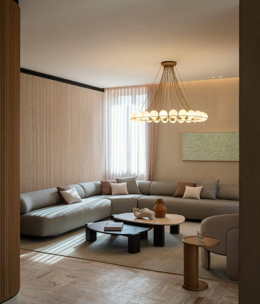 Suite living room at Six Senses Rome with a large grey sectional sofa and dark low tables, a ring chandelier of glass globes, pale cocciopesto plaster walls and a sheer-curtained window