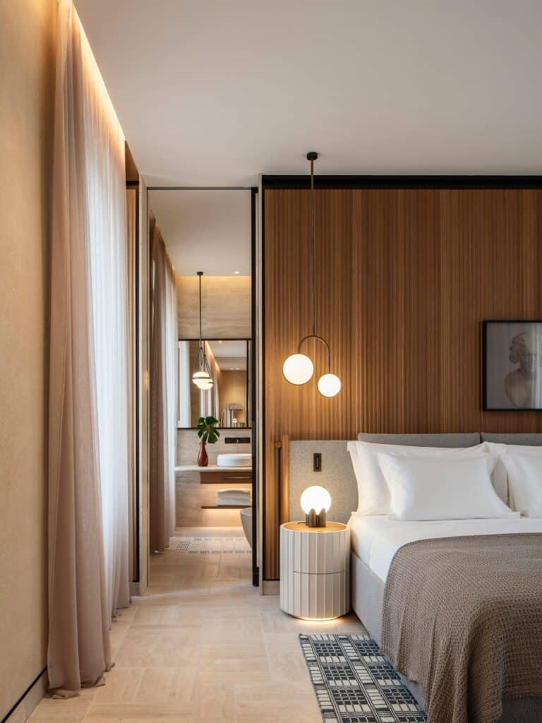 Bedroom at Six Senses Rome with a bed against a ribbed timber headboard wall, a globe pendant and lamp on a fluted bedside table, a graphic black and white rug, travertine floor and long linen curtains