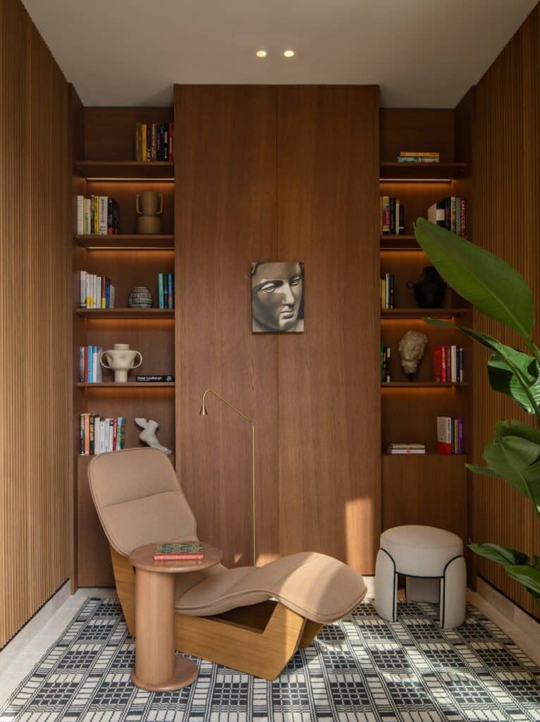 Reading corner in a Six Senses Rome suite, a tan leather lounge chair and a small round stool on a black and white patterned rug before timber shelves of books and objects with a sculpted classical face