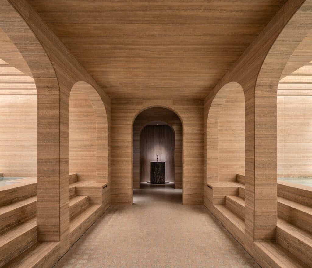 Roman baths in the spa of Six Senses Rome, a travertine-clad passage of arched openings with stepped stone seating on each side leading to a dark niche with a small stone basin