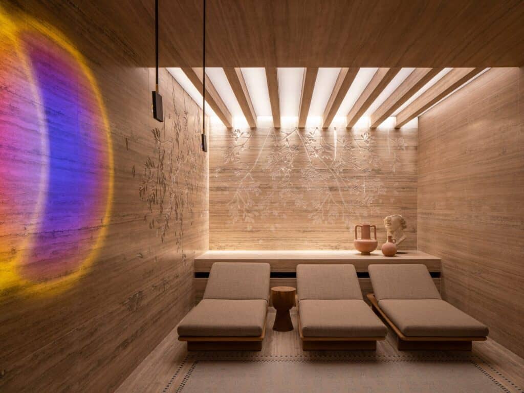 Relaxation room in the Six Senses Spa Rome with three upholstered loungers under backlit timber slats, travertine walls carved with leaf reliefs and a glowing purple and amber light artwork
