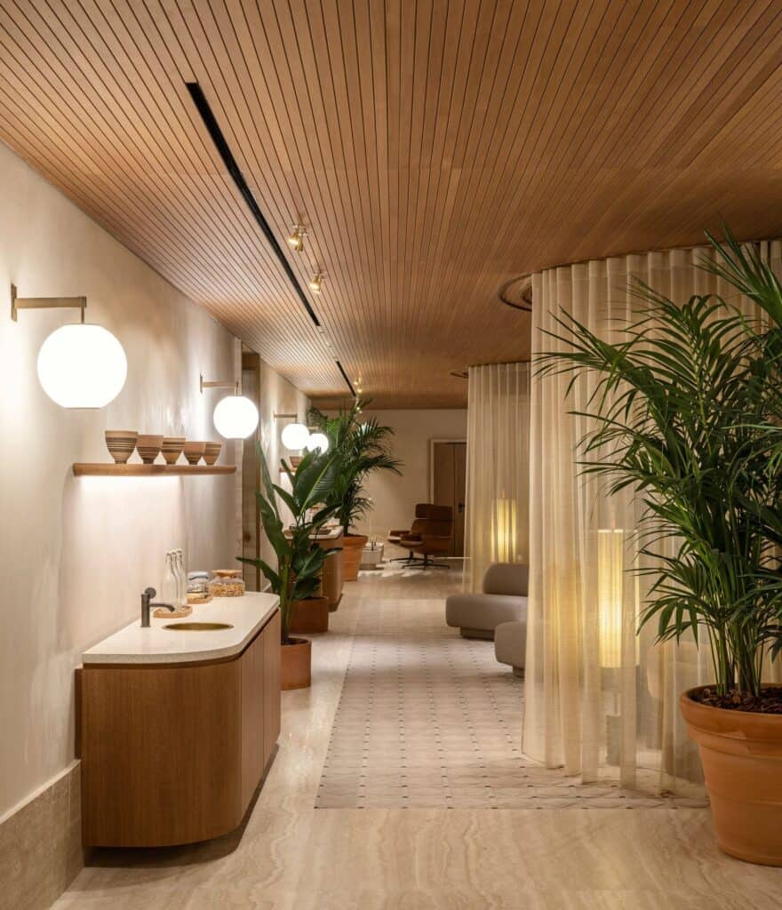 Spa corridor at Six Senses Rome under a slatted oak ceiling, with globe wall lights, a curved timber counter, sheer curtains around seating and potted palms on travertine floors