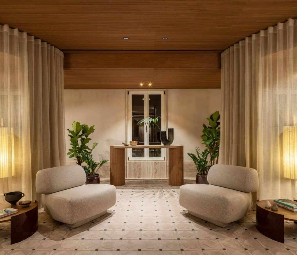 Spa waiting room at Six Senses Rome with two cream bouclé armchairs facing each other between sheer curtains, a timber console beneath a window and a patterned stone floor