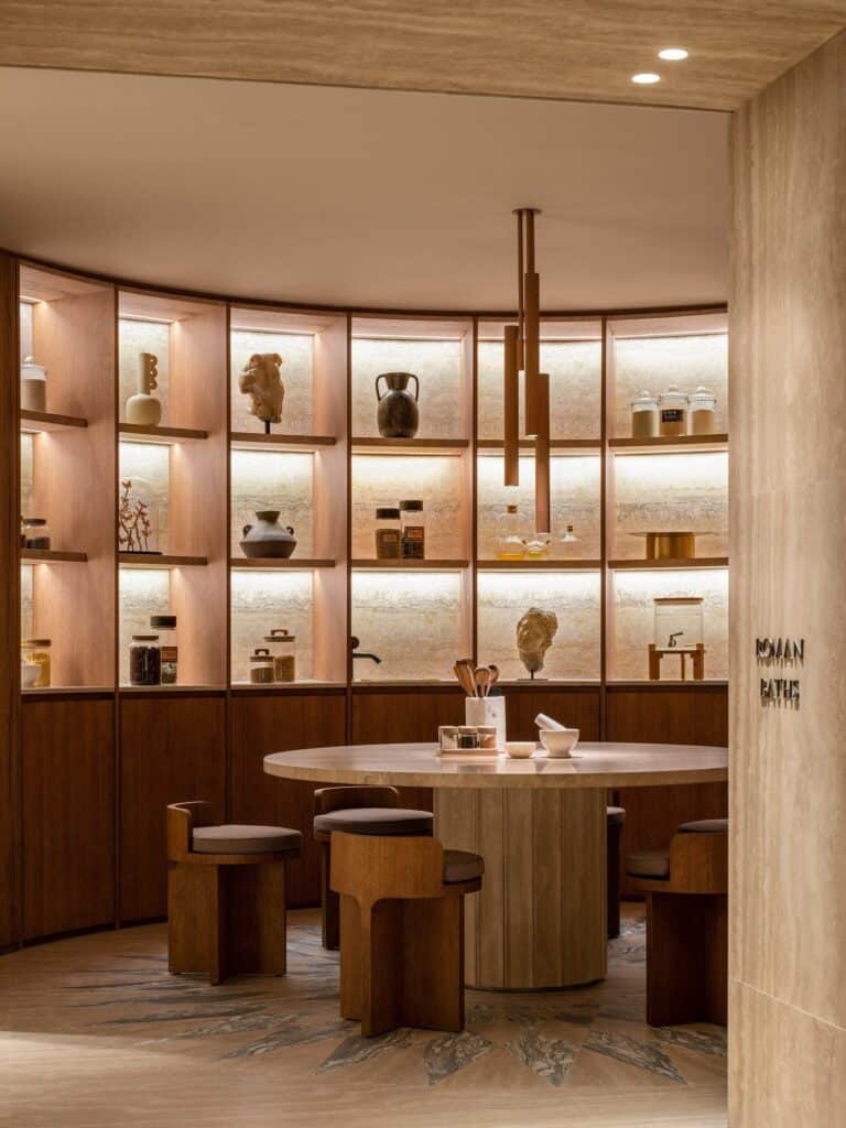 Spa tea room at the entrance to the Roman baths, a round travertine table with timber stools set in a curved niche of backlit shelves holding ceramic jars, vessels and classical busts