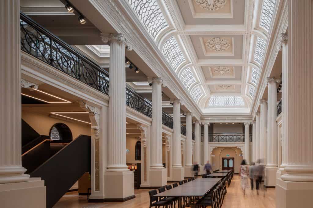 The Ian Potter Queen's Hall at State Library Victoria, restored white classical columns and iron-balustraded galleries beneath a skylit coffered ceiling, with long reading tables on a pale timber floor and a black stair to the mezzanine