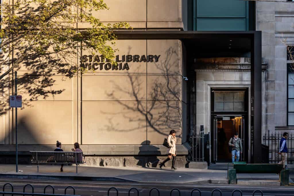 Sandstone street entrance of State Library Victoria with bronze STATE LIBRARY VICTORIA lettering, tree shadows on the wall at golden hour, people on the pavement and a black canopy over the glazed doorway
