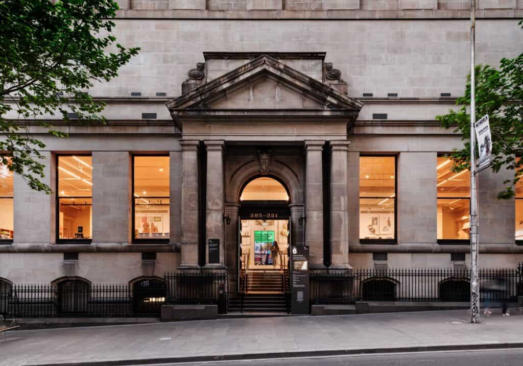 Reopened Russell Street entrance of State Library Victoria at dusk, a classical stone portico with a pediment and arched doorway between tall lit windows behind black iron railings