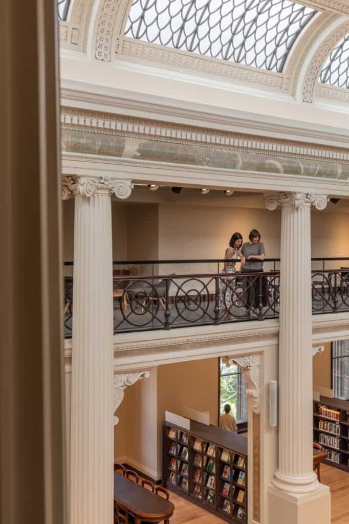 Portrait view across the restored Queen's Hall at State Library Victoria, two visitors at the iron balustrade of the mezzanine between Ionic columns, glazed skylights above and bookshelves below