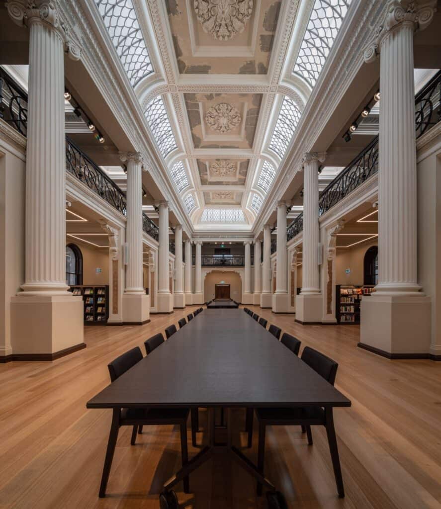 Long dark reading table with black chairs running down the centre of the Queen's Hall at State Library Victoria, framed by tall white columns, mezzanine balconies and the ornate skylit ceiling