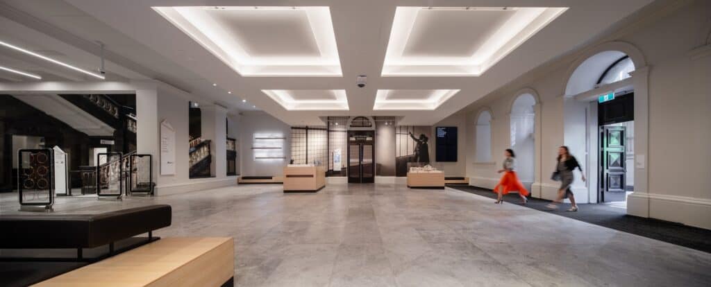 Wide panorama of a redeveloped hall at State Library Victoria with a pale stone floor, glowing recessed ceiling coffers, arched doorways and display cases, two visitors hurrying across
