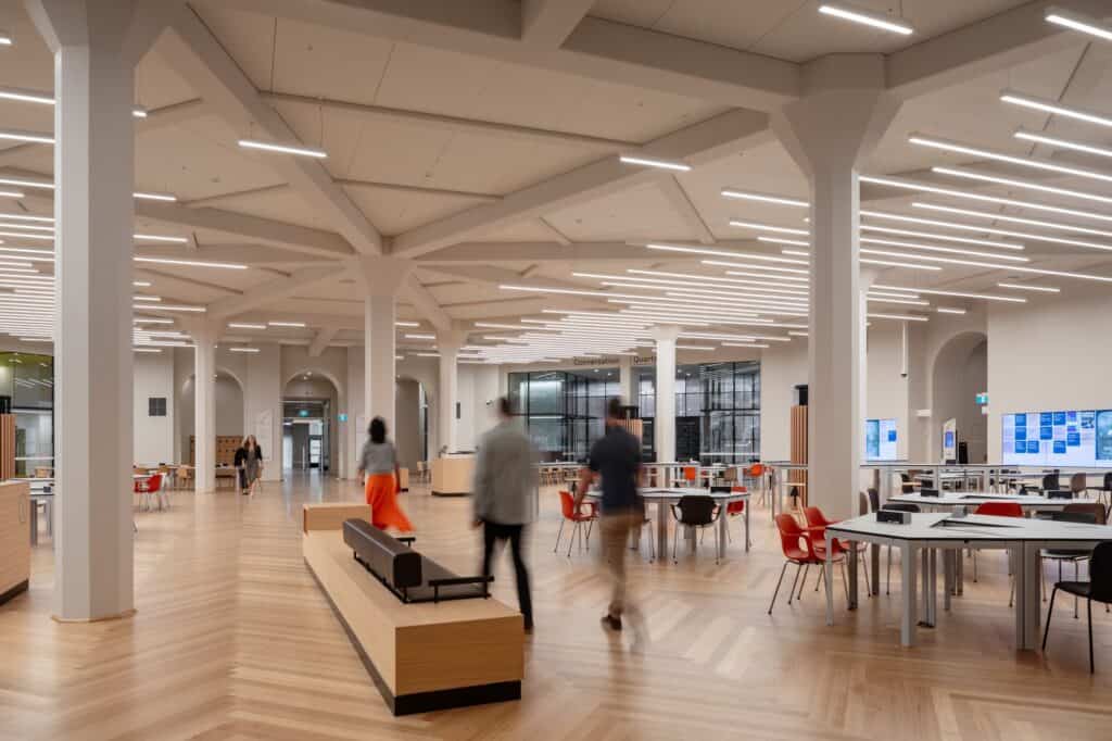 Open-plan learning space at State Library Victoria with white columns branching into a beamed ceiling, linear lights, herringbone timber floor, red chairs at long tables and a timber bench with people walking past