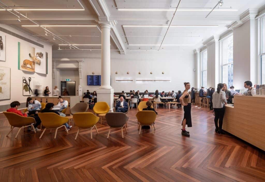 Lounge at State Library Victoria with mustard and grey armchairs on a red-brown herringbone timber floor, framed natural history prints on the wall and a timber service counter beside tall windows