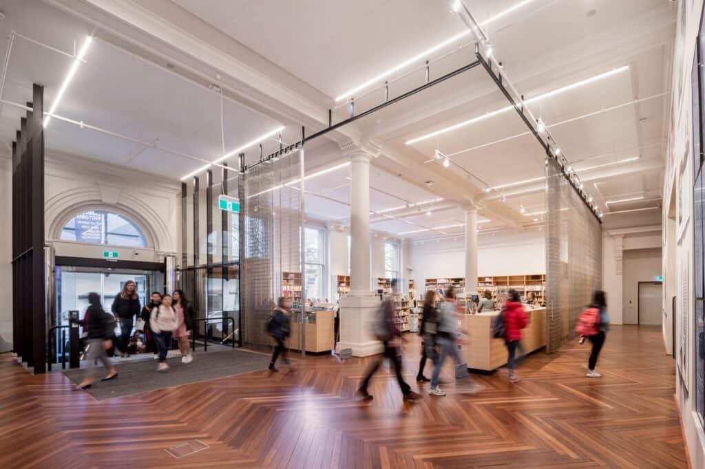 Bright welcome hall at State Library Victoria with an arched fanlight window above glass doors, a black mesh screen dividing the space, slender white columns and visitors crossing a herringbone floor
