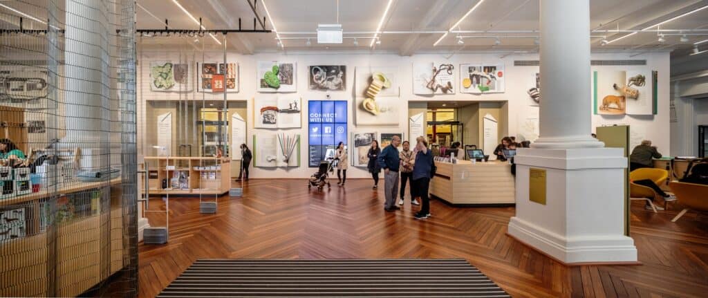 Gallery and information area at State Library Victoria with collection artworks on a white wall, a square white column, a timber counter and visitors on a herringbone timber floor beyond a wire mesh screen