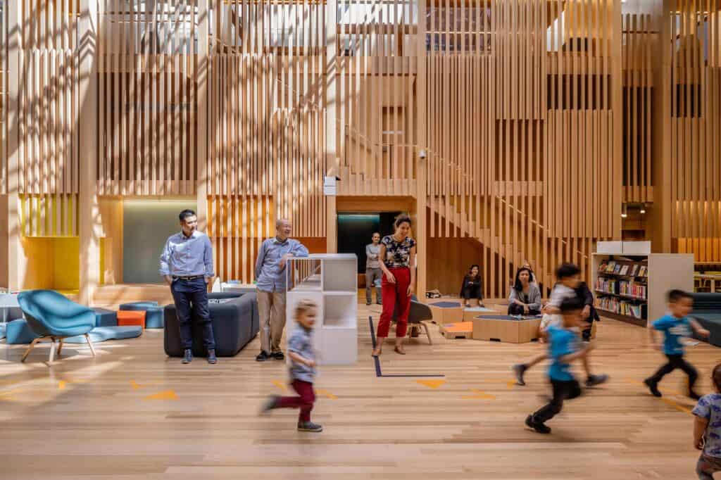 Two-level storybook castle in the Pauline Gandel Children's Quarter at State Library Victoria, built of pale vertical timber battens with a stair inside, children running across the floor past blue and navy seating